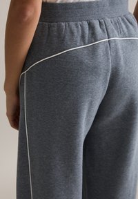 Szare spodnie dresowe z prążkowanym paskiem w talii, luźnym krojem oraz białym przeszyciem wzdłuż bocznych szwów dla kontrastu. Miękki, teksturowany materiał.