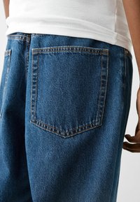 Tmavě modré džíny z denimu s kapsou na zadní straně a kontrastním prošitím a měkkým, texturovaným povrchem, doplněné bílým žebrovaným topem.