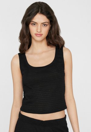 Młoda kobieta z brązowymi włosami do ramion, ubrana w czarny, teksturowany krótki top bez rękawów i pasujące spodnie, zwrócona przodem.