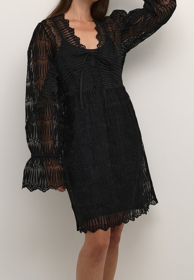 Femme portant une robe noire en dentelle transparente avec de longues manches bouffantes et des bords festonnés, debout devant un fond clair uni.