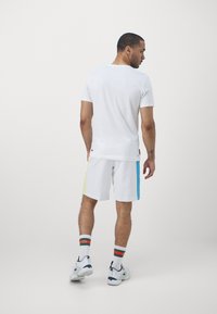 Vit T-shirt och vita shorts med blå och gula sidopaneler, i kombination med sportstrumpor och sneakers. Slätt tyg och avslappnad passform.