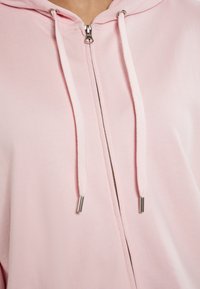 Sudadera con cremallera rosa, hecha de una suave mezcla de algodón, presenta una cremallera frontal, tela texturizada y cordones en contraste con puntas de metal.