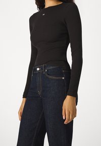 Černý dlouhý rukáv ribbovaný top s kulatým výstřihem, se malým logem na hrudi, spárovaný s tmavě modrými džínovými kalhotami s širokými nohavicemi.