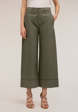 Kobieta ubrana w oliwkowe, szerokie i krótkie spodnie, zestawione z sandałami na wysokim obcasie z paskami i wzorem skóry węża oraz w białą bluzkę włożoną do środka.