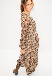 Floral Maxikleid aus durchsichtiger Stoff mit braunen, blauen und cremefarbenen Mustern. Mit einer taillierten Taille und langen Ärmeln. Getragen mit braunen Stiefeln.