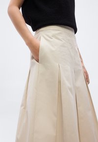 Μπεζ πλισέ φούστα από λείο ύφασμα, με πλαϊνές τσέπες και εφαρμοστό ύψος στη μέση. Το τοπ είναι μαύρο και με υφή, αποκαλύπτοντας ένα χέρι.