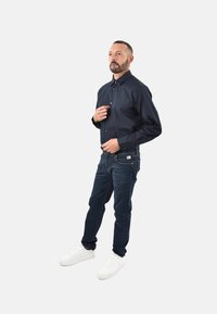 Uomo che abbottona una camicia a maniche lunghe blu navy, indossando jeans blu scuri e sneakers bianche, in piedi contro uno sfondo chiaro e uniforme.