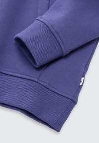 Tessuto di felpa viola con polsini a coste, che mostra una manica piegata e un orlo cucito. Piccola etichetta con logo visibile sul lato.