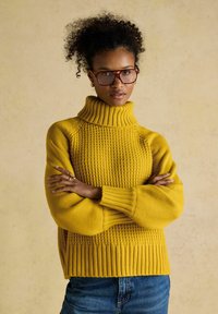 Frau mit lockigem Haar, die große Brille und einen senfgelben Rollkragenpullover trägt, steht mit verschränkten Armen vor einem neutralen Hintergrund.