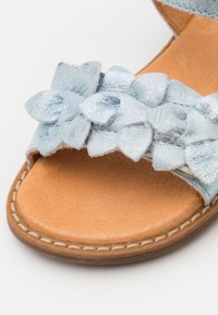 Lichtblauwe sandalen met een gestructureerde band met gelaagde satijnen strikken, een tan leren voetbed en decoratieve stiksels langs de zool.