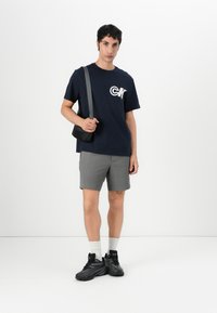 Námornícke tričko s bielym logom, šedé textúrované šortky, čierna crossbody taška a čierne športové topánky s bielymi ponožkami.