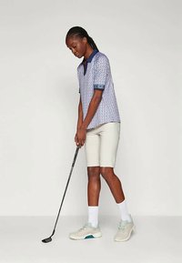 Ľahké polo tričko s vzorom v modro-bielych farbách s krátkymi rukávmi, v kombinácii s béžovými šortkami a šedými športovými topánkami, drží golfovú palicu.