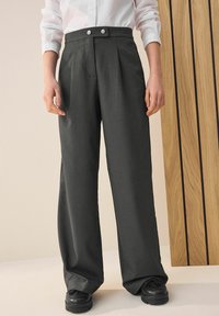 Grauweite-Hosen mit hoher Taille, ausgestattet mit zwei vorderen Falten und silbernen Knopfakzenten, kombiniert mit schwarzen, strukturierten Schuhen.