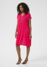 Pink short-sleeve dress with a v-neckline, tiered design, and flowing fabric. Worn with black heeled sandals. Smooth, lightweight texture.