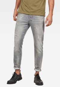 Grå denimjeans med slim fit, slitna detaljer och uppvikta fållar. Kombineras med svarta snörskor och en olivgrön t-shirt.