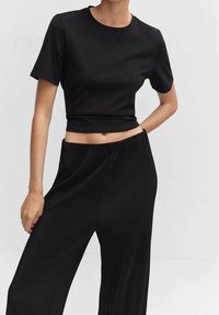 Femme portant un crop top noir ajusté à manches courtes et un pantalon noir plissé à jambes larges et taille haute, sur un fond clair uni.