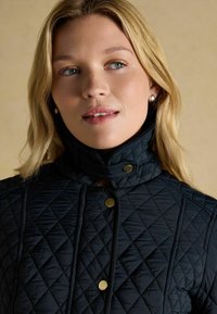 Jachetă căptușită neagră cu guler înalt, având butoane aurii și o suprafață texturată. Design simplist cu o ajustare strânsă în jurul gâtului.