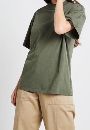 Osoba ubrana w oliwkowo-zielony t-shirt z krótkim rękawem i beżowe spodnie, z jedną ręką uniesioną blisko głowy na jednolitym białym tle.