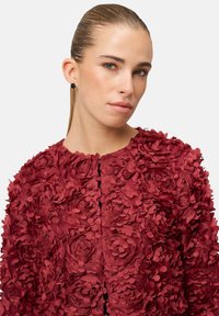 Veste bordeaux faite de fleurs en tissu superposées, design structuré, encolure ronde, fermeture éclair à l'avant, et texture florale complexe.