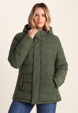 Woman with shoulder-length blonde hair wearing a green puffer jacket with a hood and blue jeans standing against a plain background.