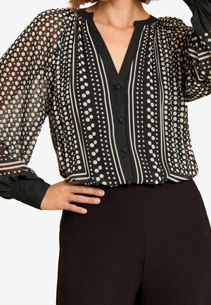 Vrouw draagt een doorschijnende zwarte blouse met witte stippen en verticale strepen, gecombineerd met een zwarte broek met hoge taille.