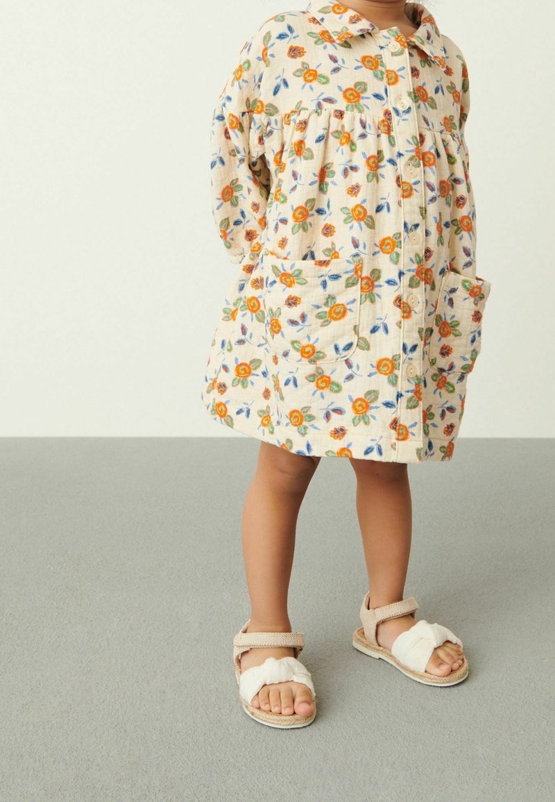 Tout-petit portant une robe crème à imprimé floral orange avec des poches frontales, associée à des sandales beige sur un sol gris.