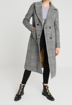 Grijze geruite trenchcoat met een grote kraag, een dubbelriem ontwerp en zijzakken, gedragen over een grijs shirt en zwarte broek. Zwarte enkellaarsjes met gouden accenten.