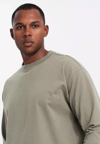 Camisa de algodón de manga larga en verde oliva con cuello redondo acanalado, que presenta una textura suave y un corte holgado. Sin patrones ni acentos visibles.
