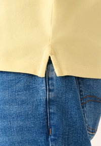 Gul bomuldsskjorte med splittet kant, kombineret med blå denimjeans med synlige syninger som en detalje.