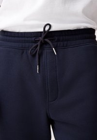 Pantalon de survêtement bleu marine avec taille élastique et cordon noir noué en nœud papillon, associé à une chemise blanche rentrée.