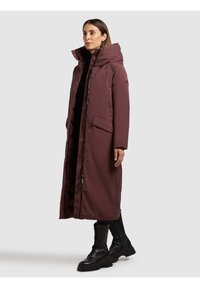 Bordó hosszú puffer kabát magas gallérral, cipzárral és patentzárakkal, oldalzsebekkel és texturált anyagból. Fekete bakancsokkal viselve.