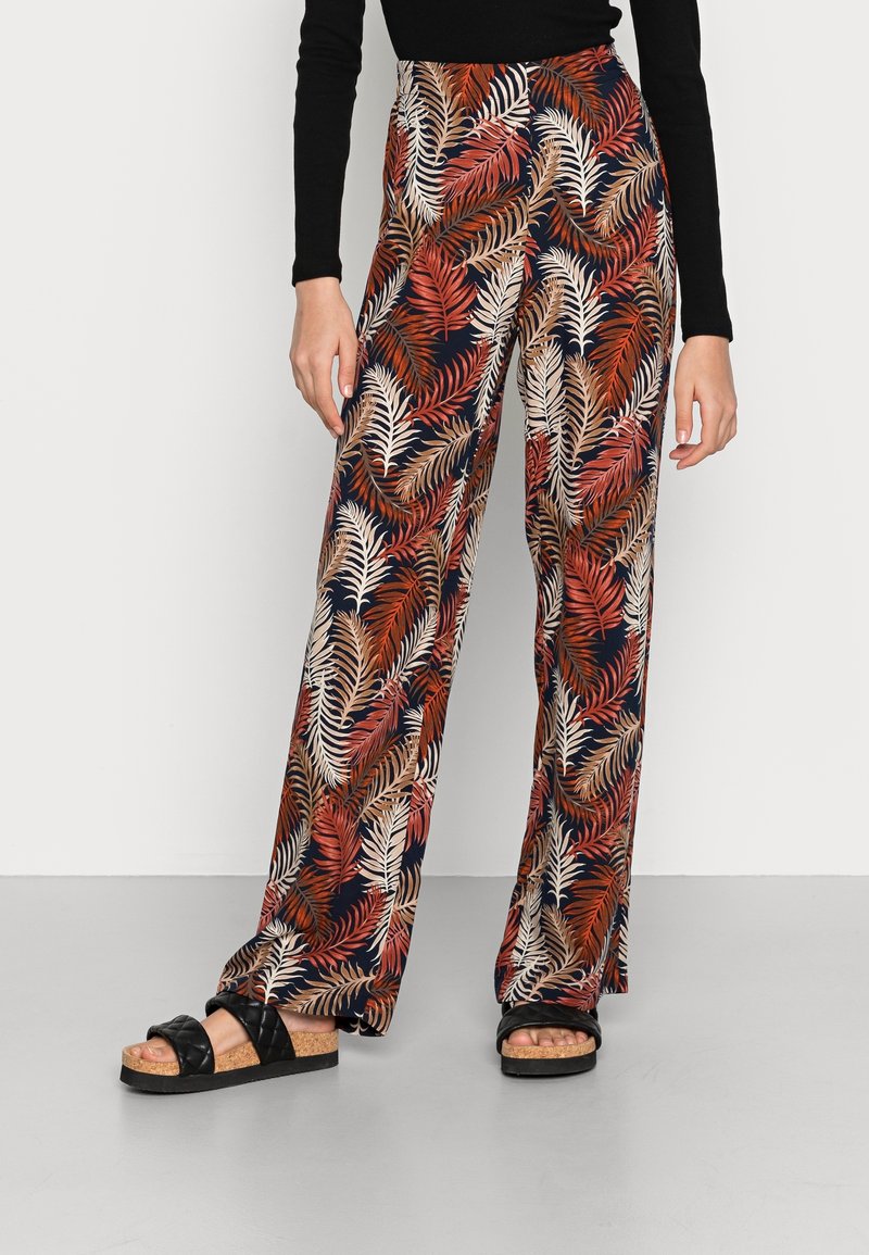 Pantalon large à motifs floraux bleu marine, avec des dessins de feuilles rouges et beiges. Matière légère avec une texture fluide. Porté avec des sandales noires.