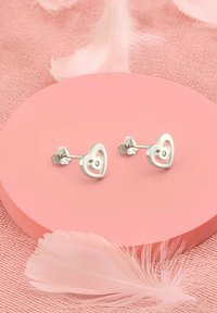 Silver hjärtformade studs med en liten central ädelsten, placerade på en rosa cirkulär yta med mjuka fjädrar som detaljer.