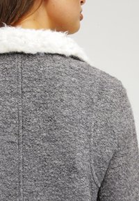 Grå texturerad kappa med en vit, fluffig krage. Tyget verkar varmt och tjockt, med en strukturerad design och synliga söm detaljer.