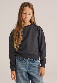 Felpa grigia scura con collo a giro e maniche a sbuffo, caratterizzata da un sottile motivo a diamante di borchie lungo il petto, abbinata a jeans blu chiaro.