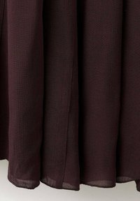 Tessuto plissettato color marrone scuro, con una texture semitrasparente, dalla forma fluida e bordi sagomati. Presenta una fessura laterale per un movimento accentuato.