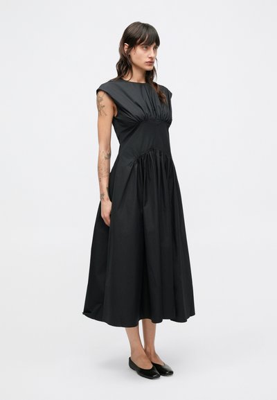 Fekete, ujjatlan ruha összehúzott felsőrésszel és lebegő szoknyával. Kerek nyakkivágással és finom vállr�észlettel rendelkezik. Sima textúrájú.