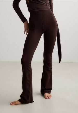 Kvinde iført brune tætsiddende flare-bukser og matchende langærmet top, stående barfodet på en lys gulv imod en ensfarvet væg.