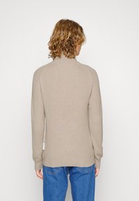 Beiger Strickpullover mit hohem Kragen, Raglanärmeln und gerippter Textur, kombiniert mit blauen Jeans. Mit einem Etikett an der Seite.