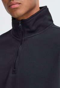 Veste en polaire noire zippée avec un col haut, présentant une texture lisse et un détail de fermeture éclair d'un quart de longueur au niveau du cou.