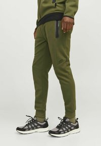 Green athletic joggers with tapered legs, featuring black zip pockets and seams, paired with black and gray mesh sneakers.
