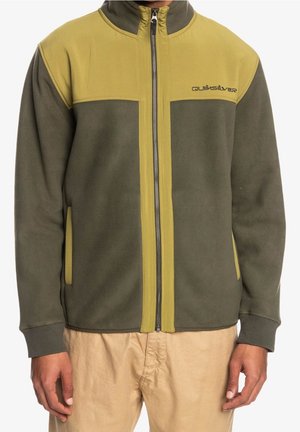Mand iført en olivengrøn og sennepsgul fleecejakke med lynlås og "Quiksilver" logo på brystet samt beige bukser, stående med arme afslappet langs siden.
