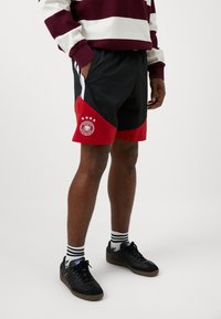 Μαύροι και κόκκινοι αθλητικοί σορτς με καμπυλωτό σχέδιο, λογότυπο με αστέρια και πλαϊνές ρίγες; κατασκευασμένοι από ελαφρύ ύφασμα.