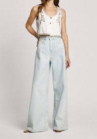 Lyhyet valkoiset toppi, jossa on koristeommelta ja puunappeja, yhdistettynä vaaleansinisiin leveälahkeisiin farkkuihin, joissa on pehmeä denimmateriaali.