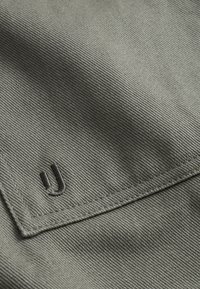 Zielony materiał o żebrowej fakturze, z wyhaftowanym złożeniem i małym, czarnym haftowanym logo "U" w rogu.