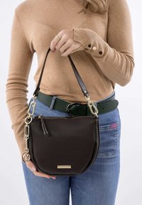 Bruine leren schoudertas met een textuurafwerking, een kettingaccent en een goudkleurige logoplaat, vastgehouden door een persoon in een vijftienkleurige trui.