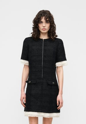 Femme portant une robe noire texturée à manches courtes avec une fermeture éclair à l'avant, deux poches avant, et un bord plissé blanc sur les manches et l'ourlet.