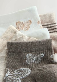 Quatre paires de chaussettes tricotées en beige, blanc et marron avec des motifs de papillons et des designs texturés, disposées en chevauchement sur un fond clair.