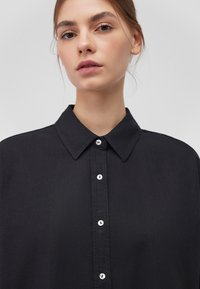 Black button-up shirt made of smooth fabric. Features a pointed collar, front placket with metal buttons, and a loose fit. No patterns or accents.