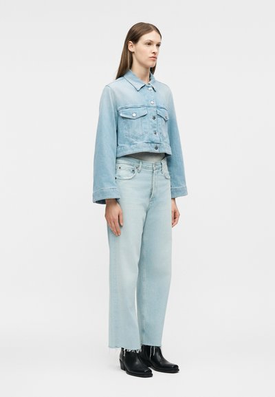 Γυναίκα που φοράει cropped τζιν μπουφάν σε ανοιχτό μπλε και φαρδιά ξεθωριασμένα τζιν παντελόνια, σε συνδυασμό με μαύρες μποτάκια σε αστράγαλο, στέκεται μπροστά σε απλό φόντο.